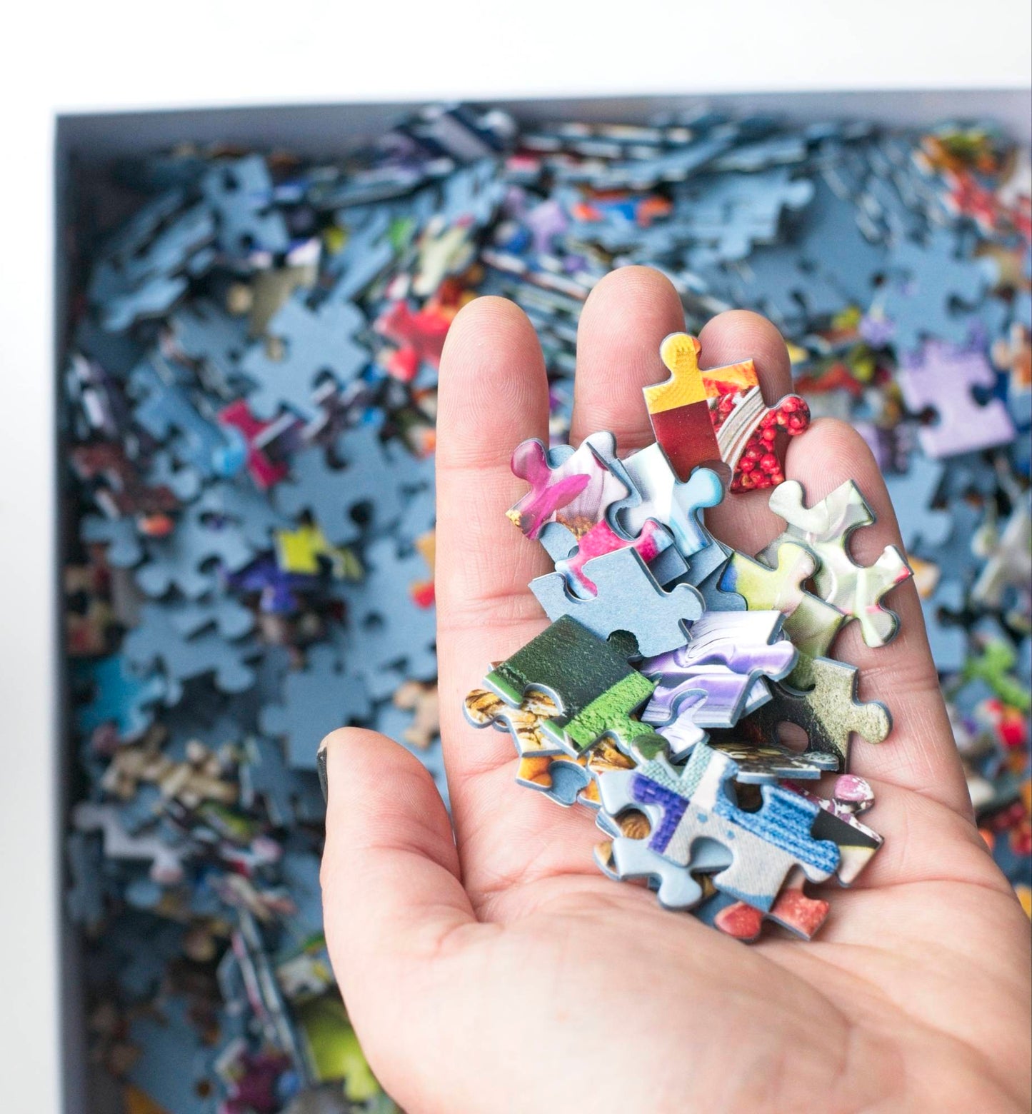 A hand holding many small colorful traditionally cut puzzle pieces made with blue board over a box filled with puzzle pieces.