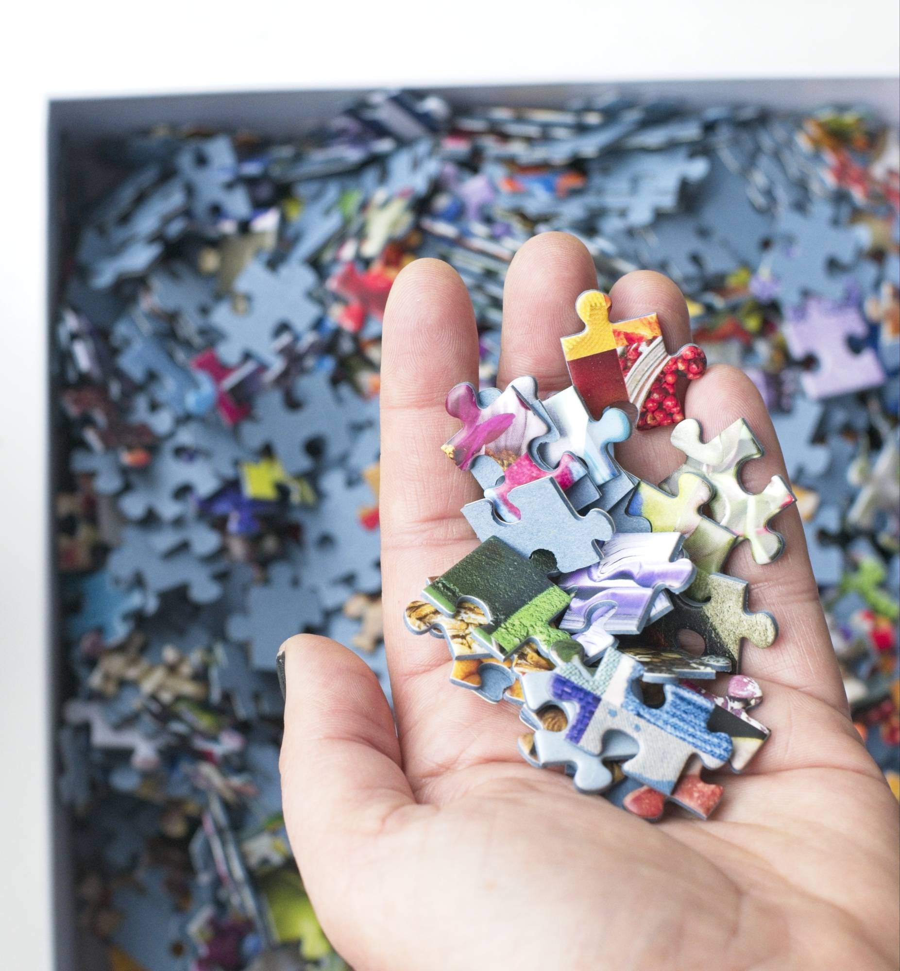 A hand holding many small colorful traditionally cut puzzle pieces made with blue board over a box filled with puzzle pieces.