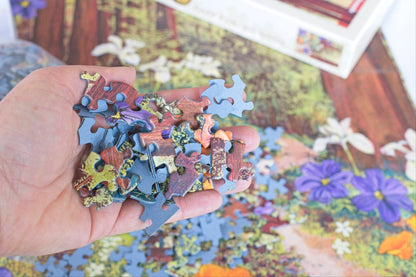 A hand holding many small brightly colored jigsaw puzzle pieces made with blue board above other scattered pieces, a poster and a product box out of focus in the background.