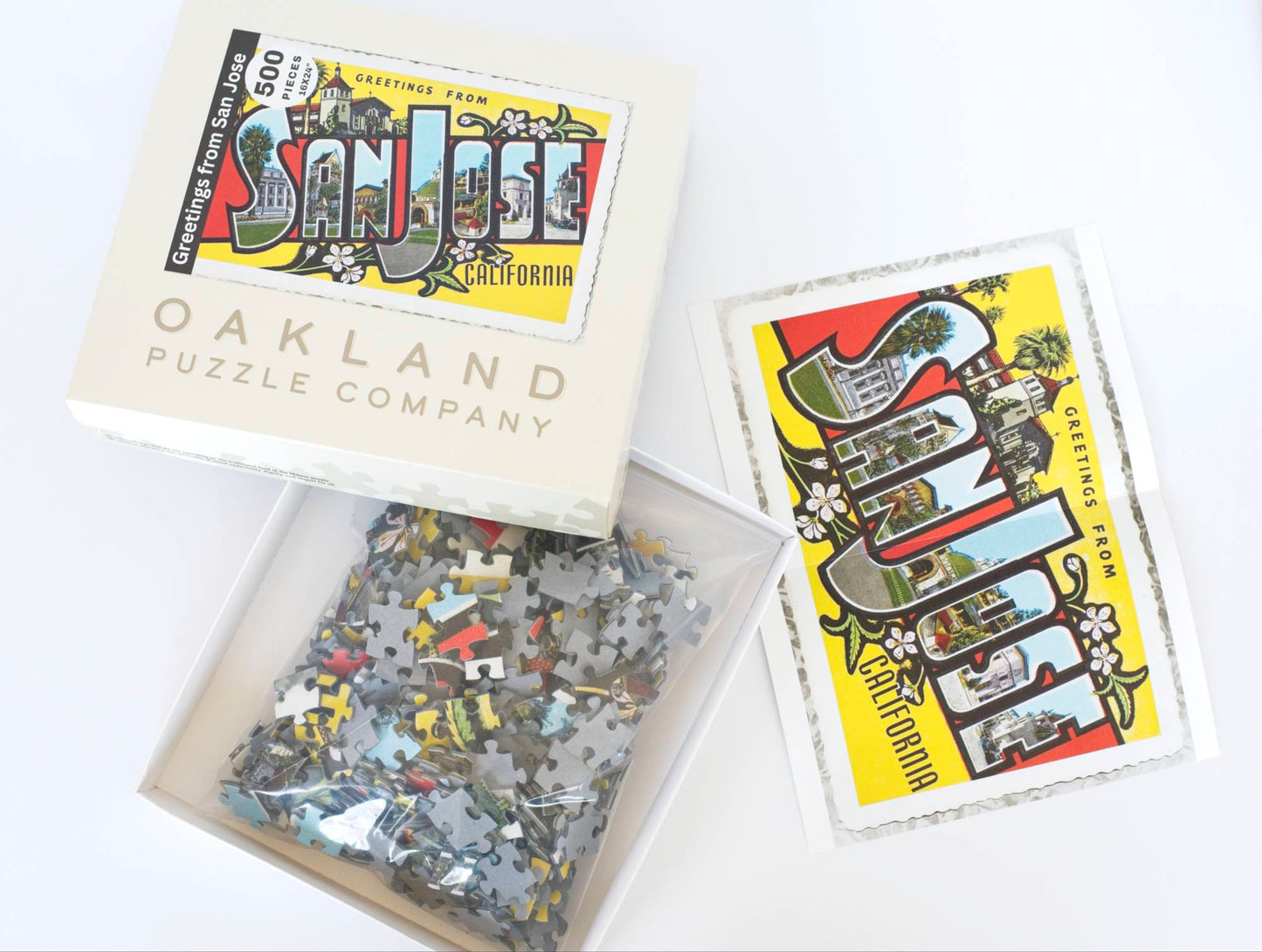 An open puzzle box labeled with the Greetings from San Jose image revealing a resealable plastic bag filled with puzzle pieces. Next to the box is a small poster and reference image for the puzzle.