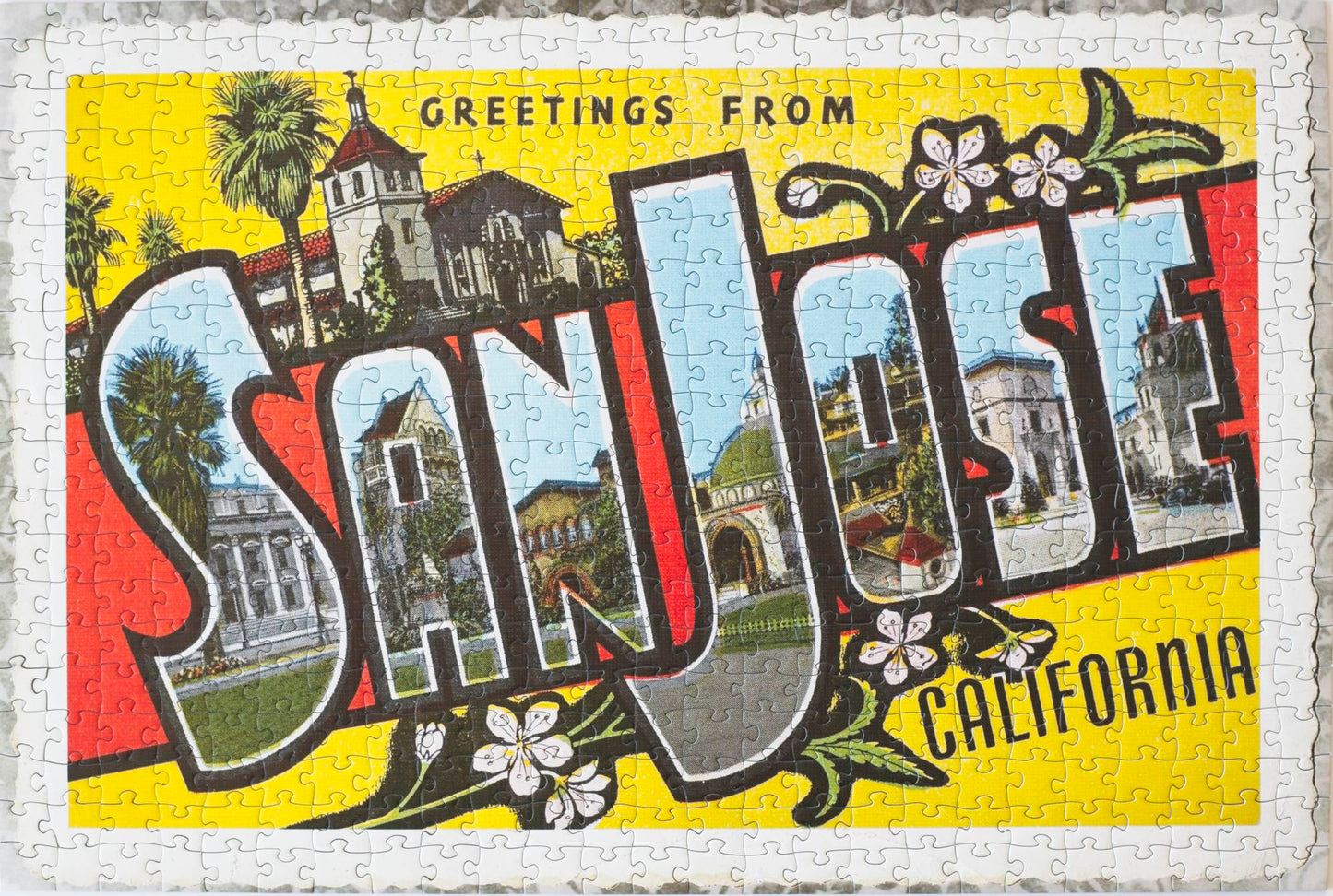 An image of a completed jigsaw puzzle depicting a vintage postcard with block letters that read Greetings from San Jose California with frayed edges laying on an old cloth. Buildings are featured in the letters of the  words San Jose.