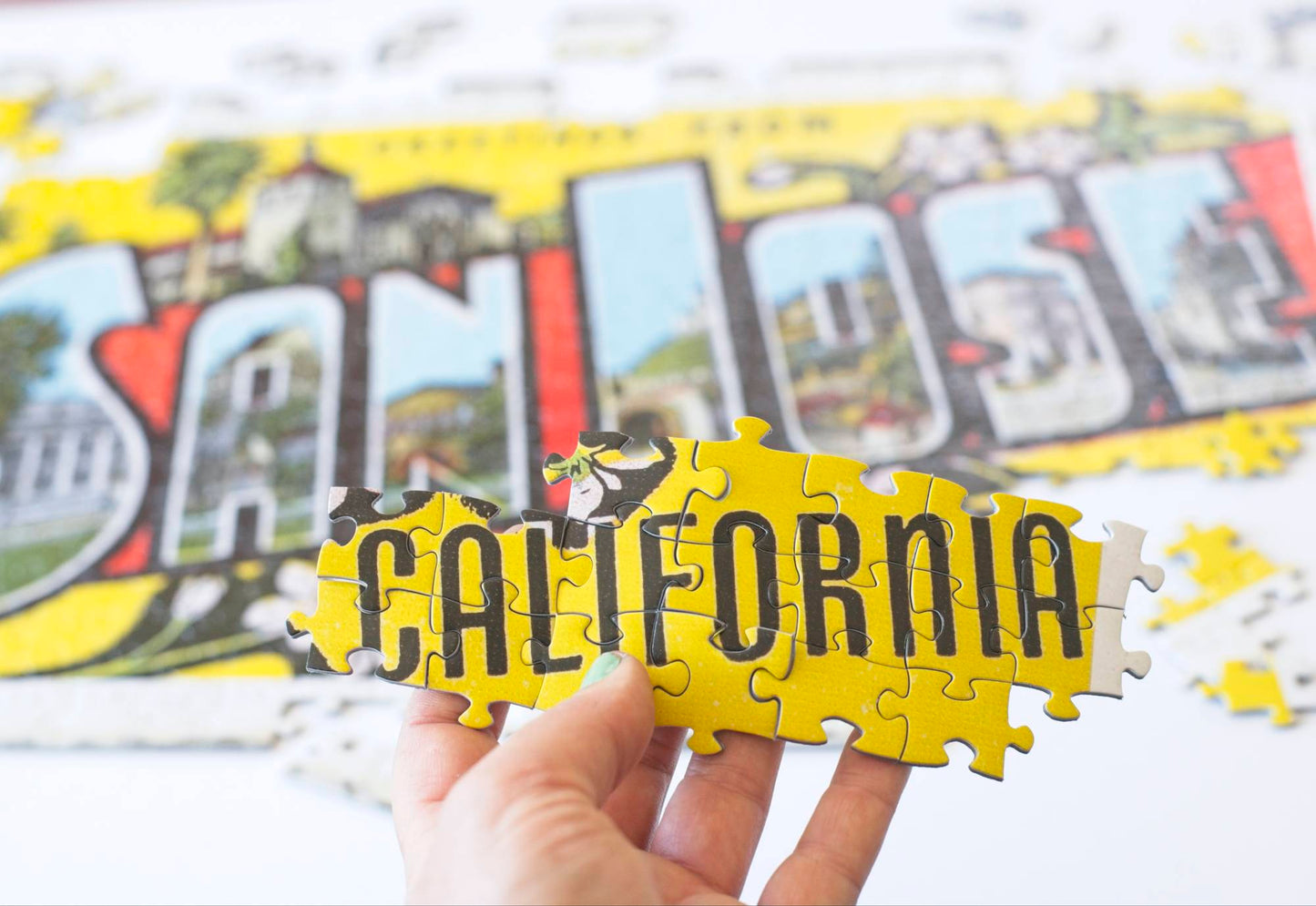 A hand holding interlocking puzzle pieces that show a yellow background with the word "California" in all caps in front of a slightly blurry partially completed jigsaw puzzle with the words San Jose.