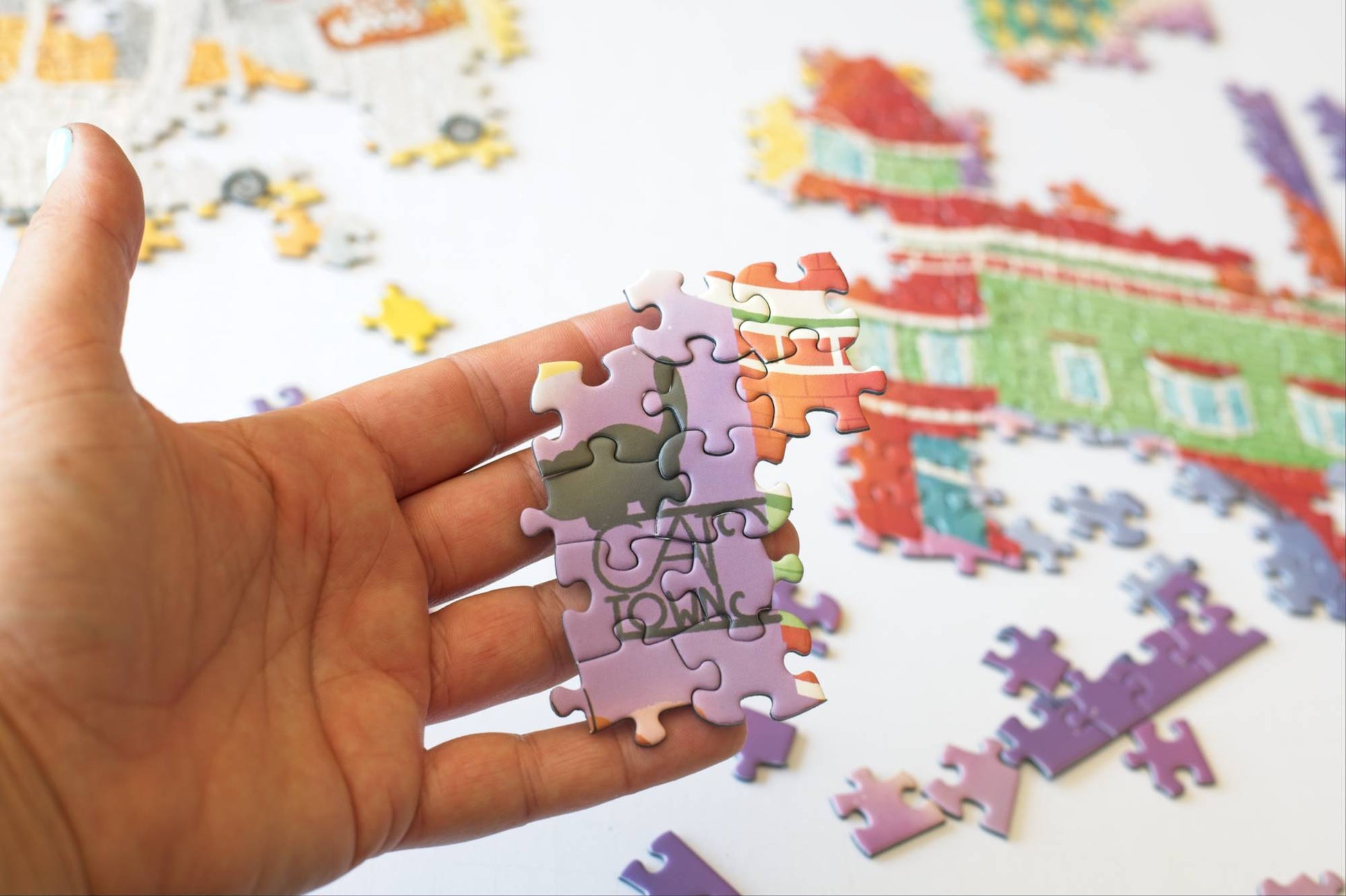 Hand holding a puzzle piece with 'Cat Town' on it, with a partially completed puzzle and scattered pieces in the background.