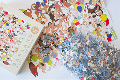 Jigsaw puzzle pieces spilling out of a resealable plastic bag onto a colorful puzzle image next to a branded Oakland Puzzle Company box