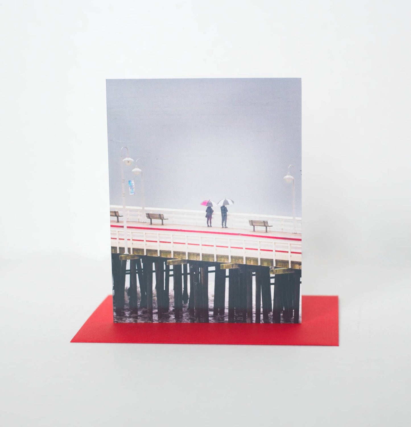 A photo of a greeting card featuring a photo of two people looking over the edge of the pier onto the grey ocean. Their umbrellas and pier detail have red and the card sits on a red envelope