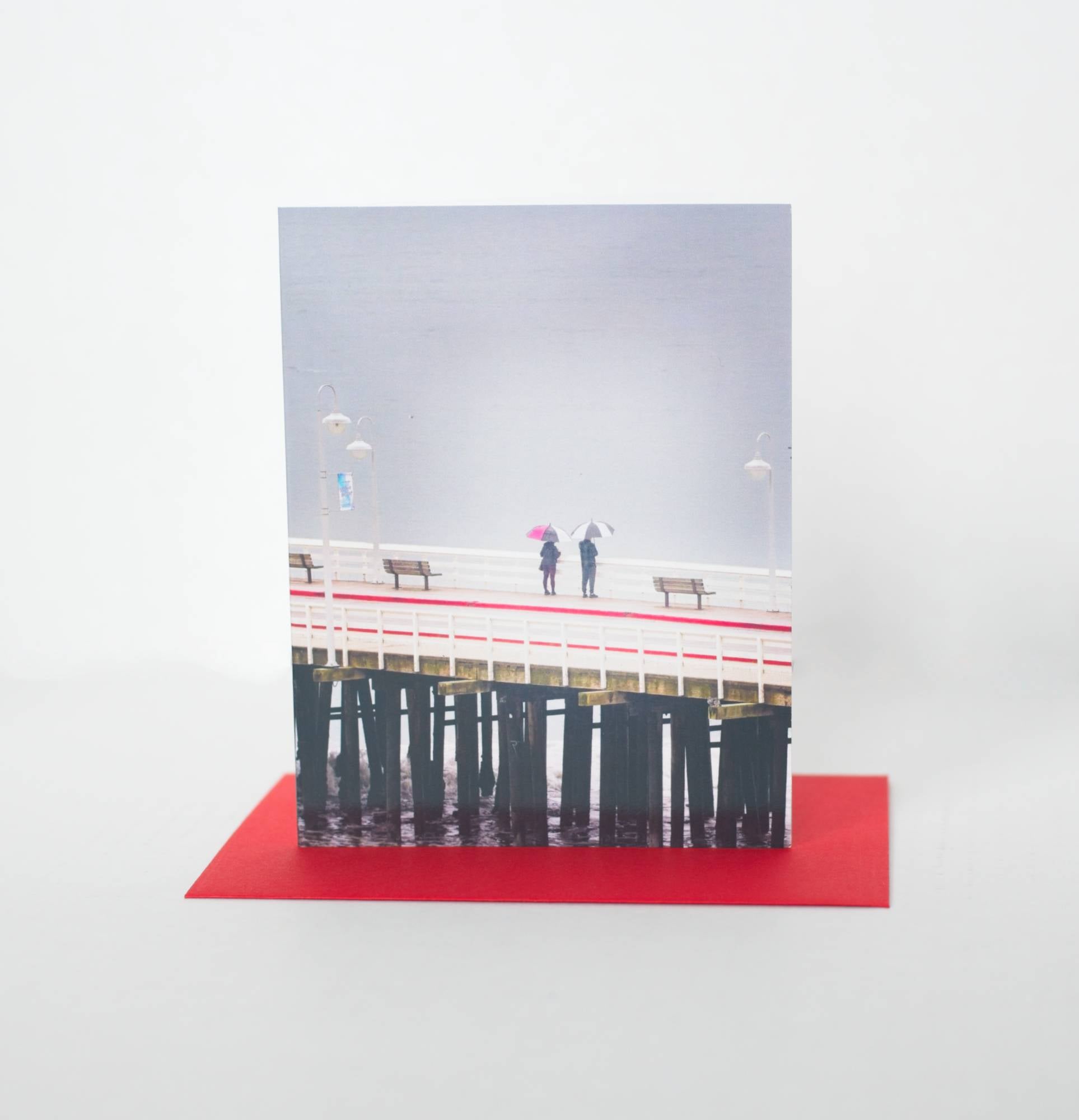 A photo of a greeting card featuring a photo of two people looking over the edge of the pier onto the grey ocean. Their umbrellas and pier detail have red and the card sits on a red envelope