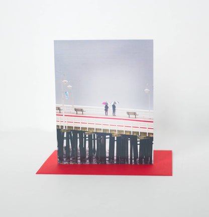 A photo of a greeting card featuring a photo of two people looking over the edge of the pier onto the grey ocean. Their umbrellas and pier detail have red and the card sits on a red envelope