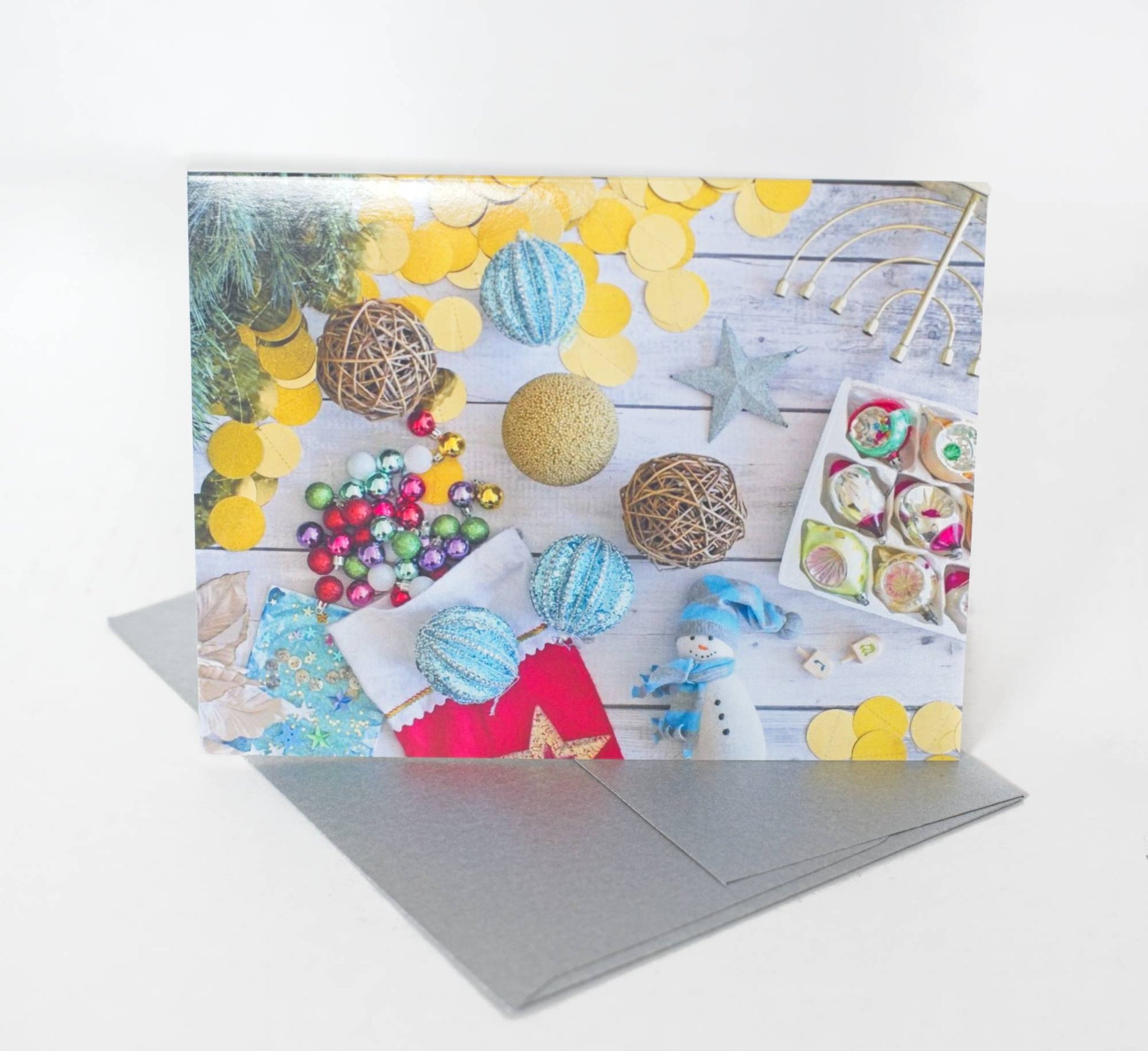 A photo of a greeting card that features a flatlay photo of a beachy rustic table scattered with ornaments and jewish and christian winter holiday items. The card sits on a silver envelope