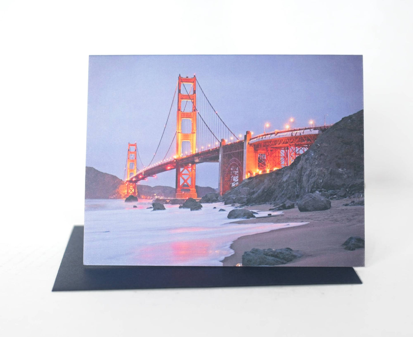 A photo of a greeting card that features a photo of the shoreline leading up to a lit golden gate bridge at blue hour. The card sits on a dark navy envelope.