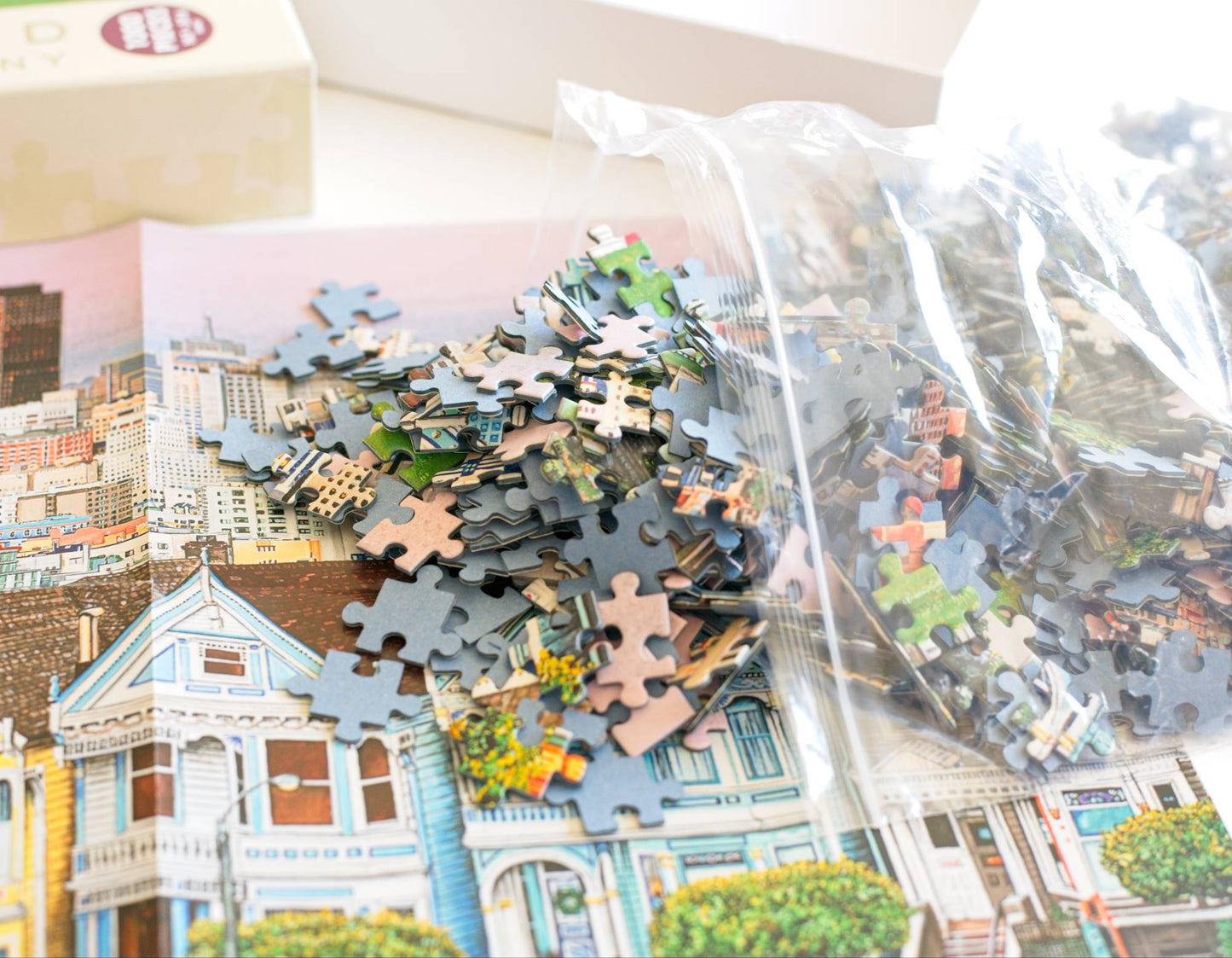 Colorful jigsaw puzzle pieces spliing out  of a resealable plastic bag onto a cityscape design next to a box.