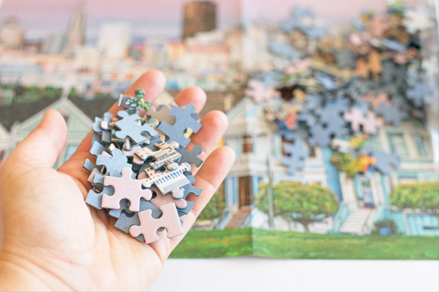 Hand holding colorful puzzle pieces with blue board backing and a completed puzzle of a cityscape in the background