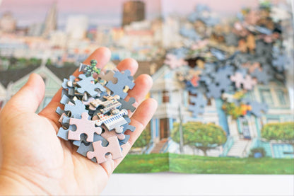 Hand holding colorful puzzle pieces with blue board backing and a completed puzzle of a cityscape in the background