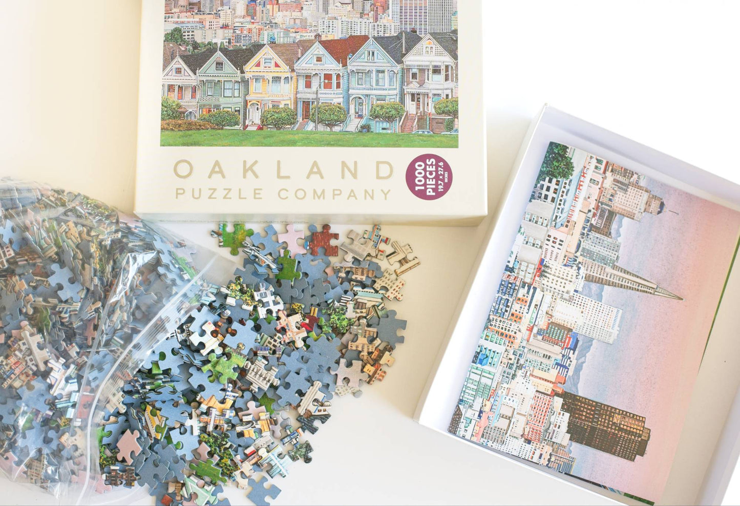 Jigsaw puzzle pieces spilling from a resealable plastic bag and the Oakland Puzzle Company packaging showing a reference image inside the open box.
