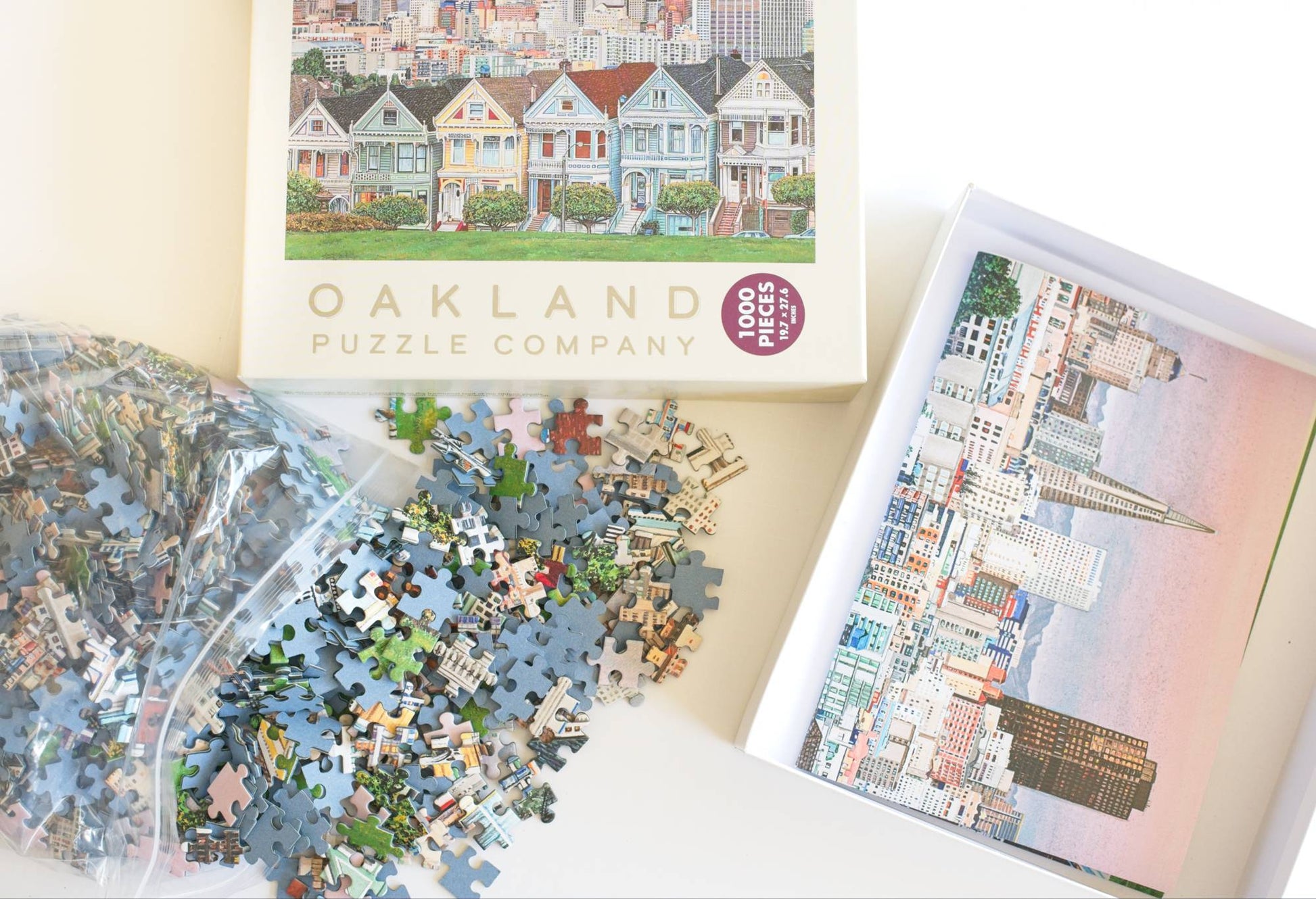 Jigsaw puzzle pieces spilling from a resealable plastic bag and the Oakland Puzzle Company packaging showing a reference image inside the open box.