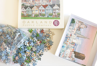 Jigsaw puzzle pieces spilling from a resealable plastic bag and the Oakland Puzzle Company packaging showing a reference image inside the open box.