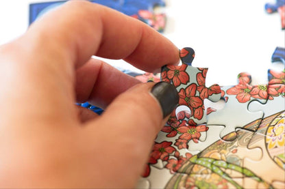 A close-up of a hand with black nail polish inserting a puzzle piece with a red flower design onto a partially assembled multicolor puzzle.