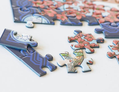 Several puzzle pieces from a jigsaw puzzle are scattered on a white surface, with a partially assembled piece visible in the background. The puzzle design features dark blue sections with light blue circuit-like patterns and pink floral sections.