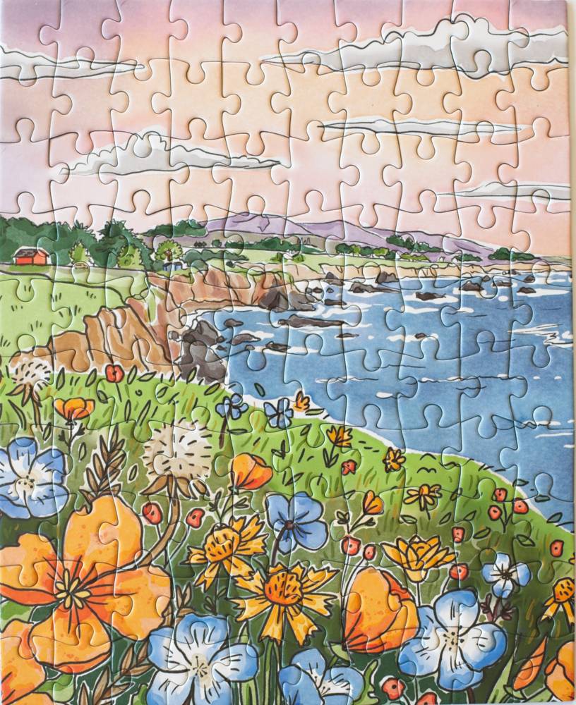 A completed jigsaw puzzle showing a colorful coastal landscape with blue water, rocky shoreline, green grass, wildflowers, and a soft pink sky with clouds.