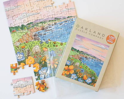 A 100-piece mini jigsaw puzzle from Oakland Puzzle Company, shown with its illustrated box with a resealable plastic bag and several loose pieces on a white surface. The puzzle features a colorful coastal landscape with flowers in the foreground.