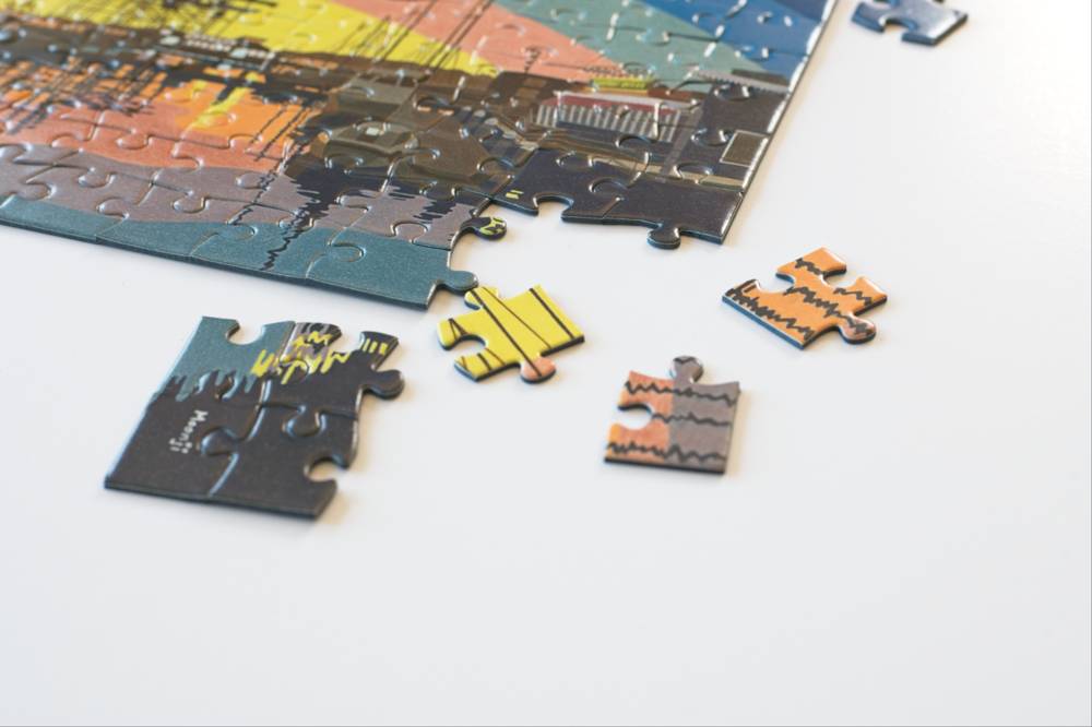 A partially completed jigsaw puzzle with several loose pieces scattered on a white surface. The puzzle shows a colorful abstract harbor landscape design with dark gray, blue, yellow, orange, and black tones.