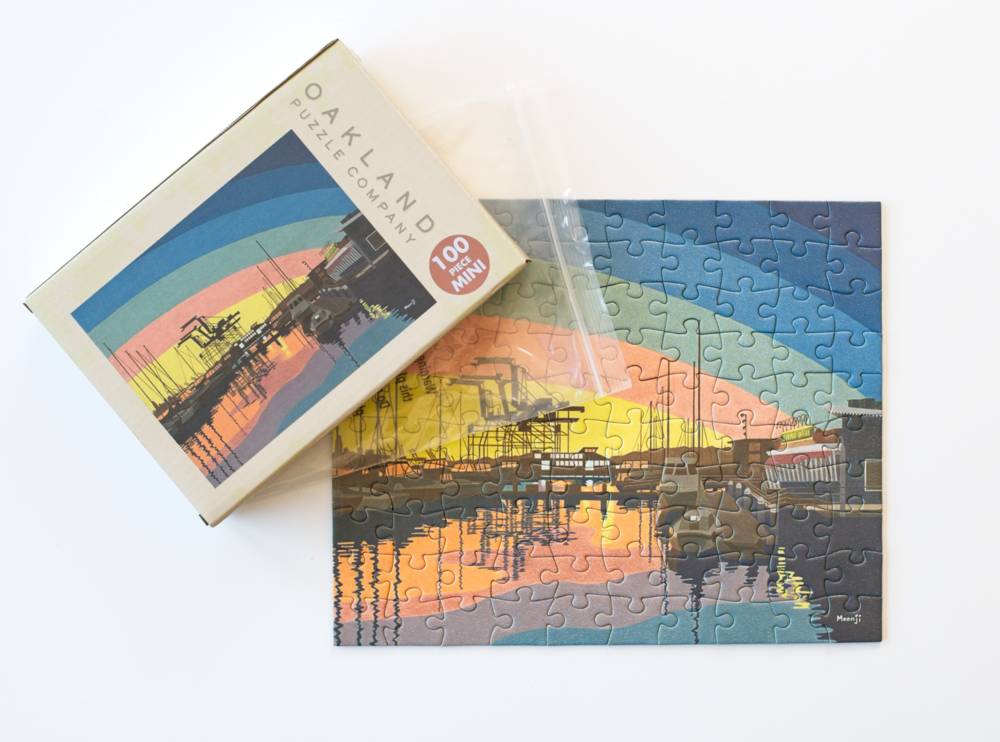 A 100-piece jigsaw puzzle and its box from the Oakland Puzzle Company, featuring a colorful sunset harbor scene with a rainbow arch over the water.