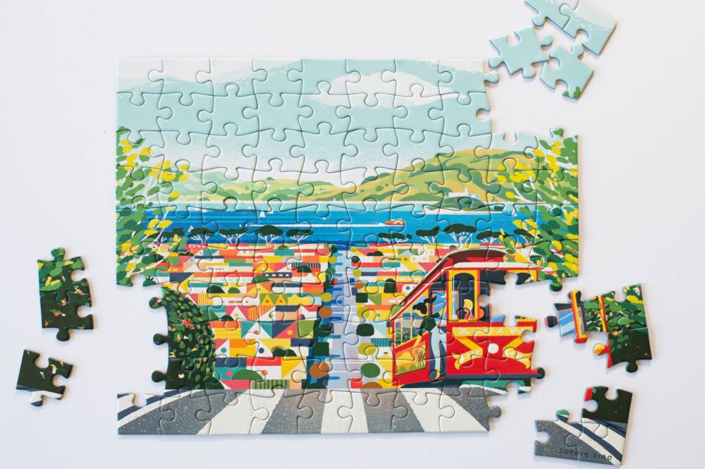 A colorful jigsaw puzzle showing a scenic coastal town with a red cable car, blue water, green hills, and a striped crosswalk, photographed on a white background with several loose puzzle pieces around it.