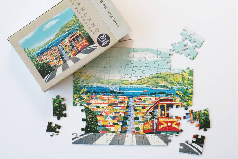 A 100-piece jigsaw puzzle featuring a cable car in a coastal scene, shown with its cardboard box, a partially assembled puzzle, and several loose pieces on a clean white background.
