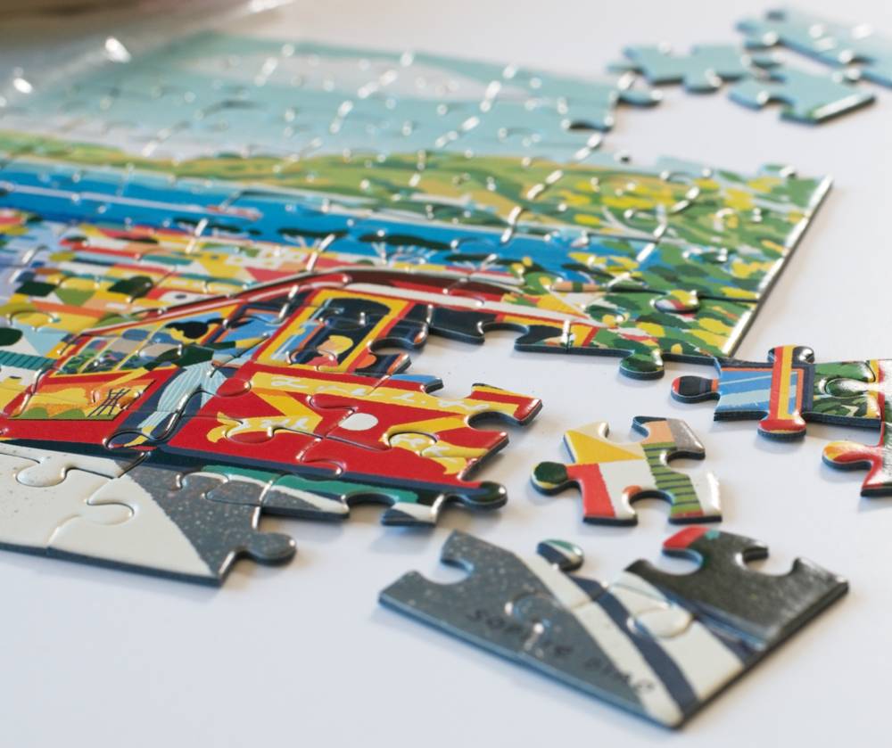 A close-up of a colorful jigsaw puzzle showing a red and yellow cable car scene, with several loose puzzle pieces scattered on a white surface.