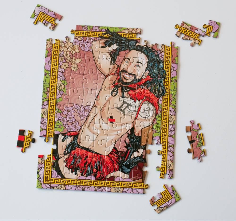 A partially assembled jigsaw puzzle featuring a stylized illustration of a shirtless bearded man with long dark hair, red feather-like accents, and a pink floral background, surrounded by loose puzzle pieces on a white surface.