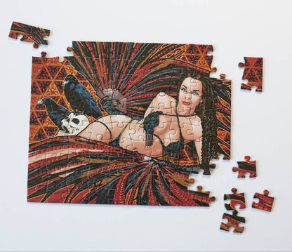 A nearly completed jigsaw puzzle with several loose pieces on a plain white background. The puzzle artwork shows a reclining woman in a black bikini surrounded by bold red, orange, and black feather-like patterns, with a blue bird and a skull visible.