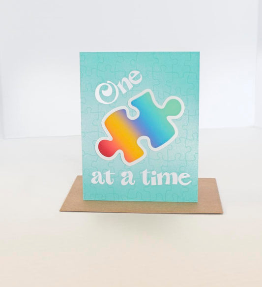 A greeting card with a jigsaw puzzle background and a rainbow sticker shaped like a puzzle piece. Text on the card says One 🧩 at a time 