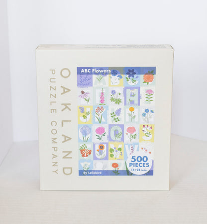 A jigsaw puzzle product box showing a pastel grid image of flowers over the alphabet with Oakand Puzzle Company branding.