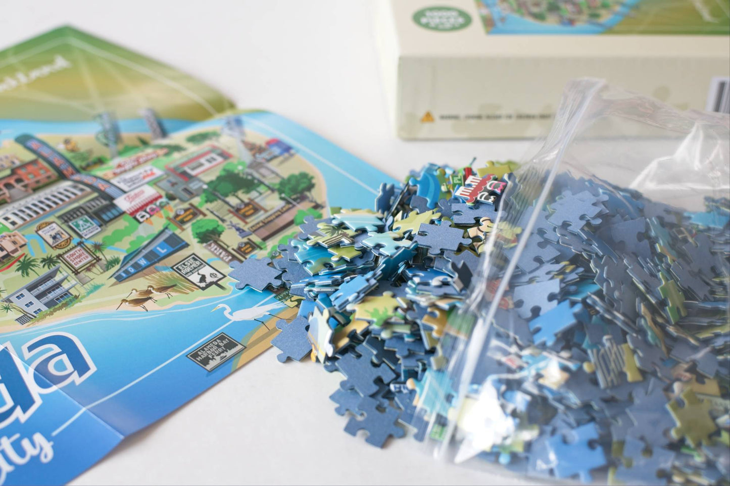 A close-up of a jigsaw puzzle showing loose blue puzzle pieces spilled from a resealable plastic bag, with part of the illustrated puzzle box and printed poster visible on a white surface.