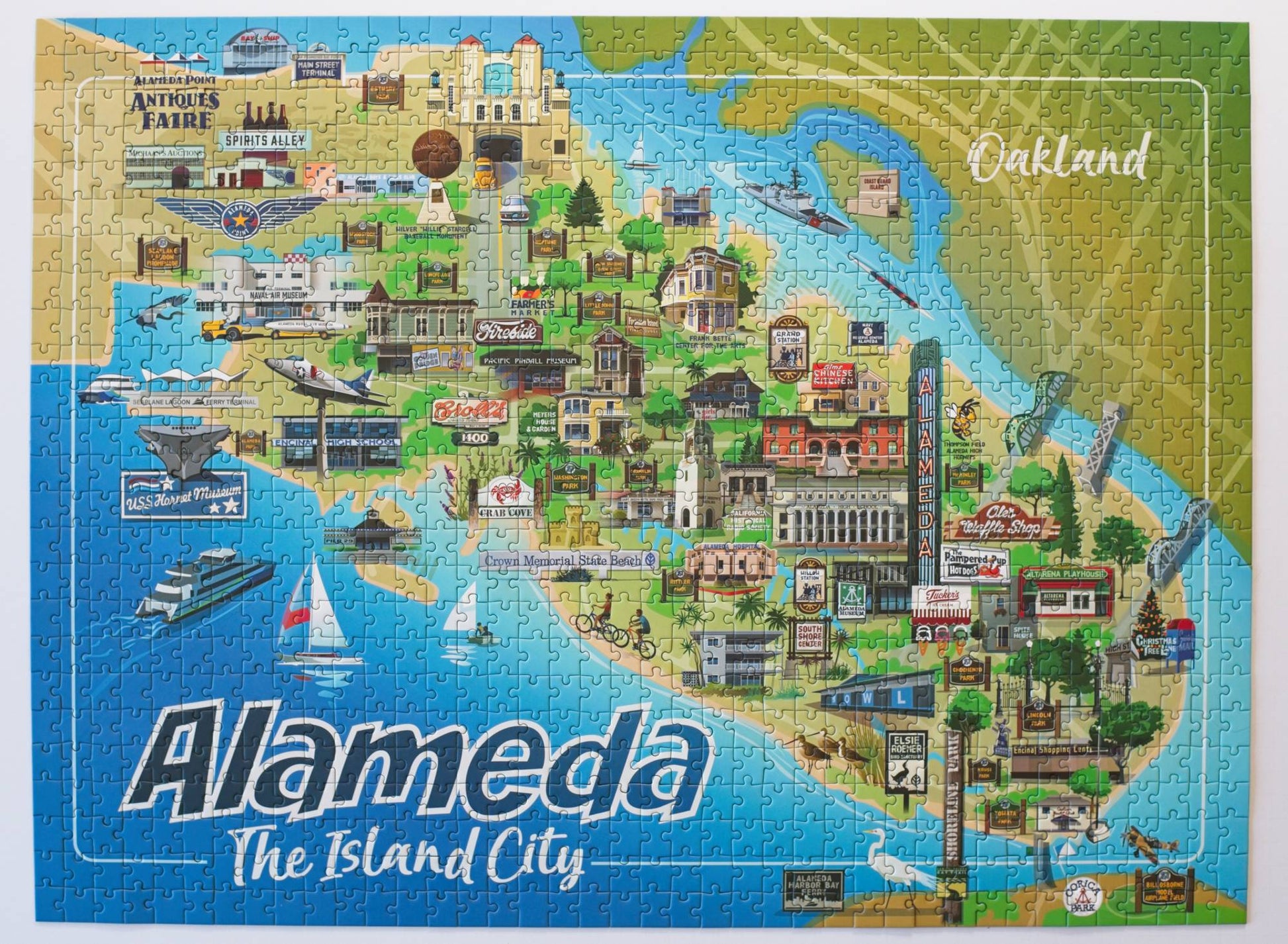 A completed jigsaw puzzle featuring a colorful illustrated map of Alameda, California, with the words 'Alameda The Island City' at the bottom and 'Oakland' in the upper right.