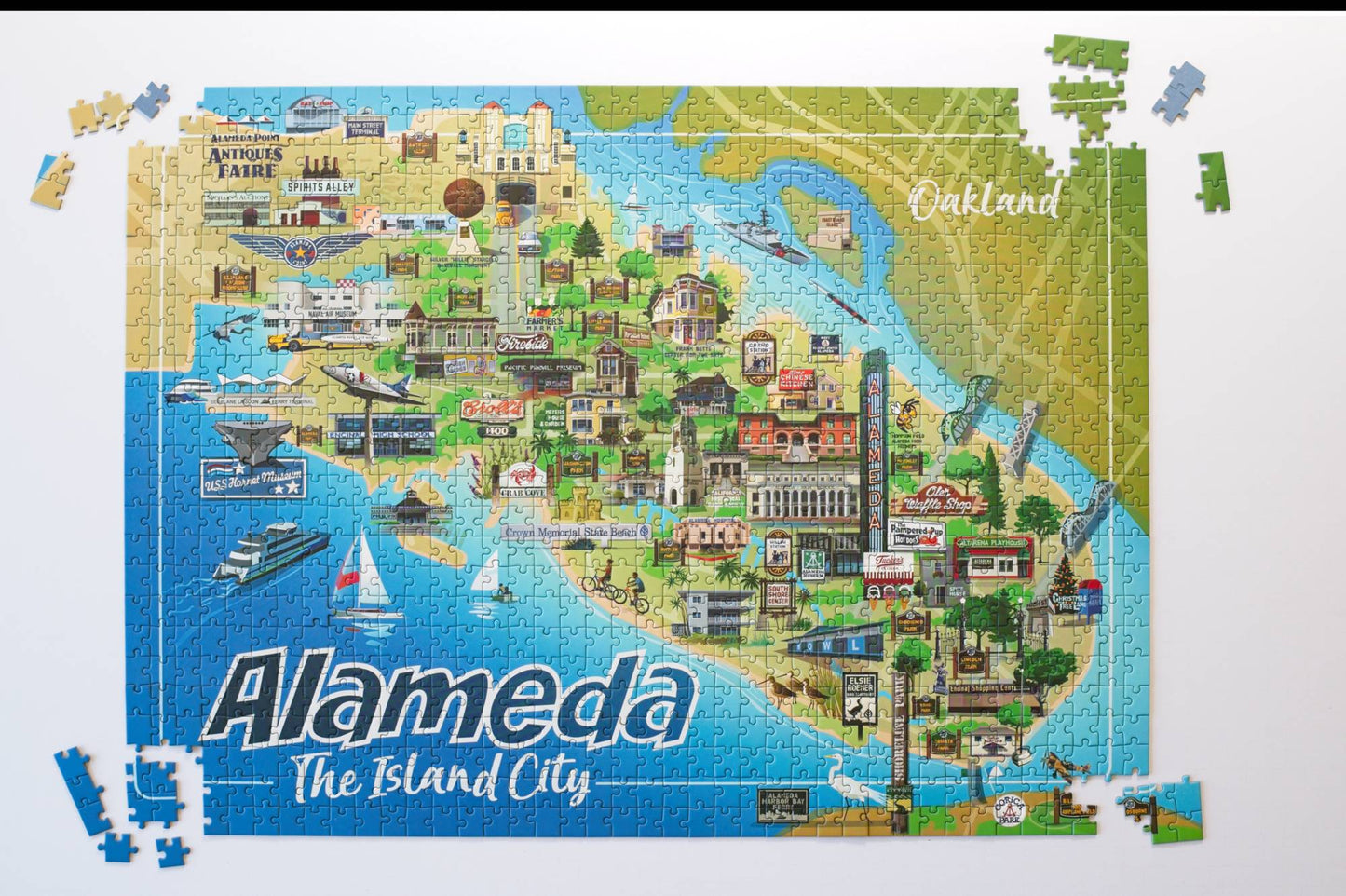 A completed illustrated jigsaw puzzle of Alameda, California, showing a colorful city map with water, boats, buildings, and the text 'Alameda The Island City.' Several loose puzzle pieces are scattered around the edges on a white background.