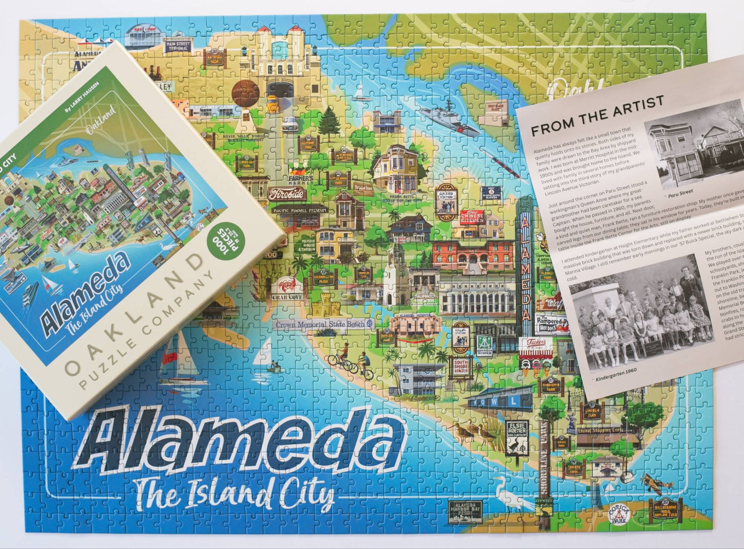 An Alameda-themed jigsaw puzzle showing a colorful illustrated map of the city, with the puzzle box, a note from the Artist, layered on the completed puzzle on a white background.