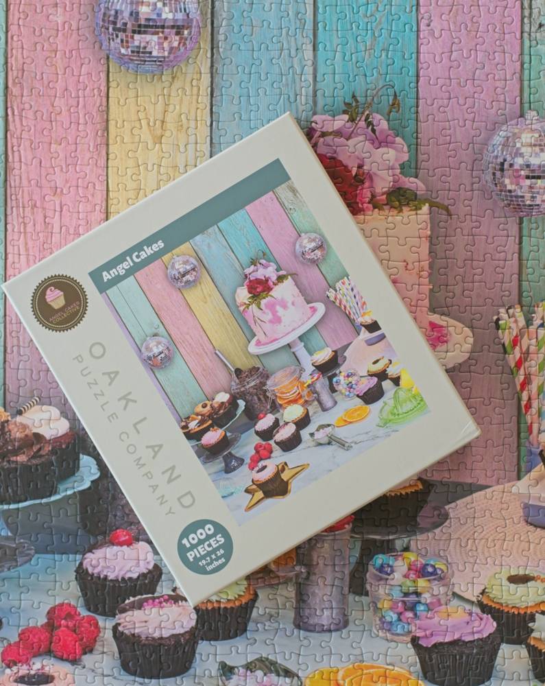 Oakland Puzzle Company 'Angel Cakes' 1000-piece jigsaw puzzle box, displayed diagonally on a colorful, themed background of pastel wooden planks, cupcakes, and party decorations.