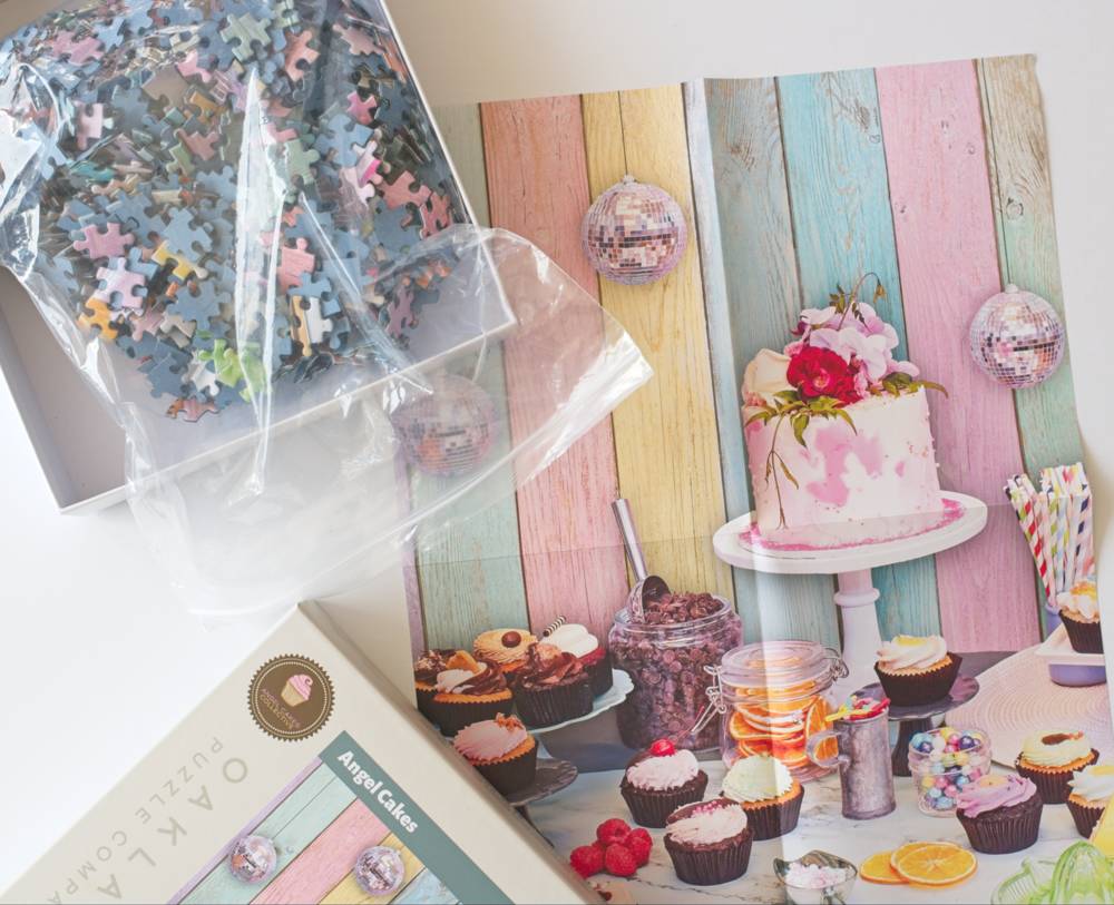 The Angel Cakes jigsaw puzzle box and packaging. The design features colorful wooden planks in pink, blue, yellow, and green with cupcakes, a pink cake, and decorative sweets.