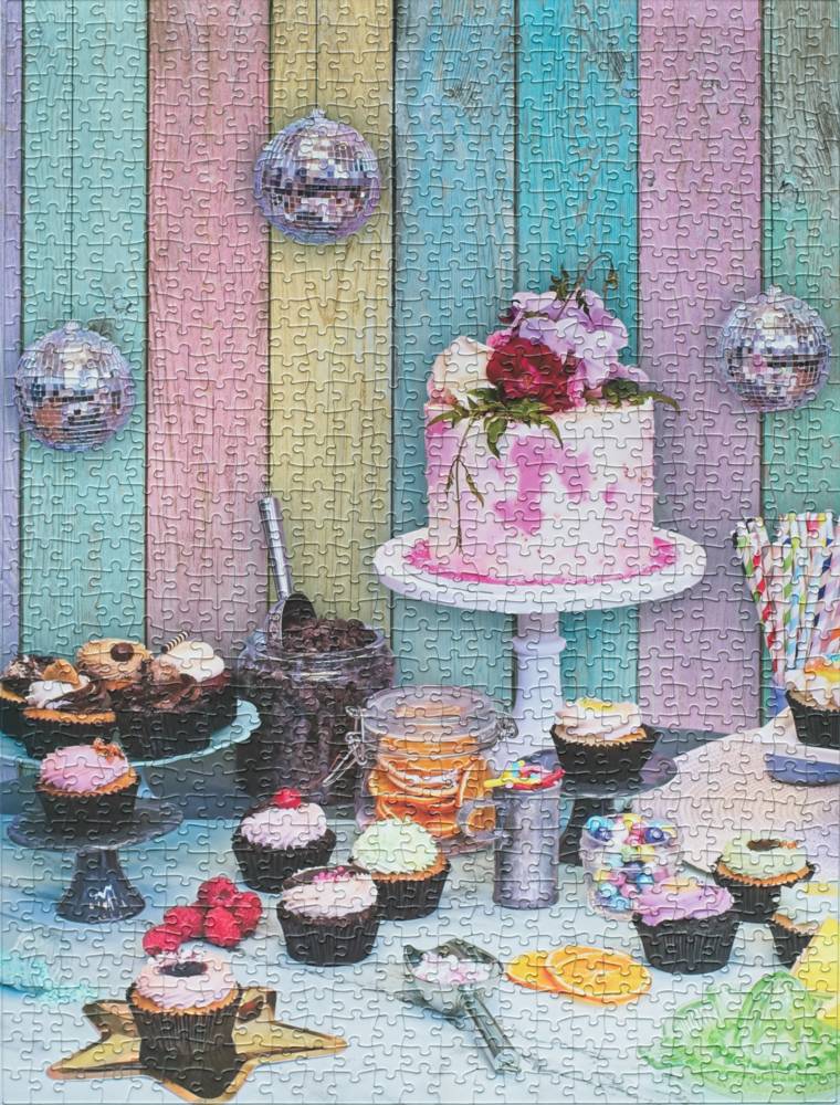 A colorful jigsaw puzzle showing a party table setup with a pink frosted cake on a white pedestal, cupcakes, candy, and disco ball decorations against a pastel wooden plank background.