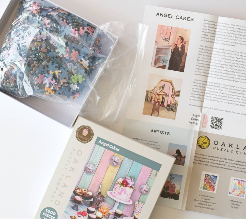 An open puzzle box showing the included contents: a bag of multicolored puzzle pieces, the open Angel Cakes poster with photos and text, and the front cover of the Oakland Puzzle Company box, on a clean white background.
