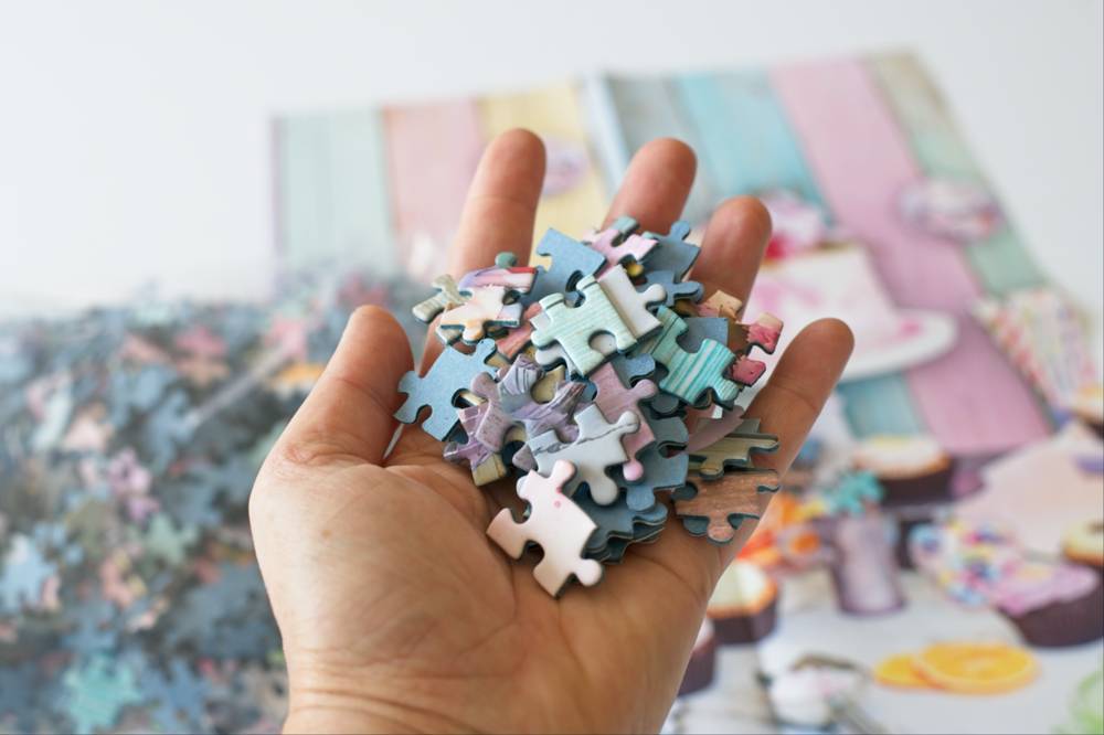 A hand holding a pile of pastel-colored jigsaw puzzle pieces, with the puzzle packaging blurred in the background.