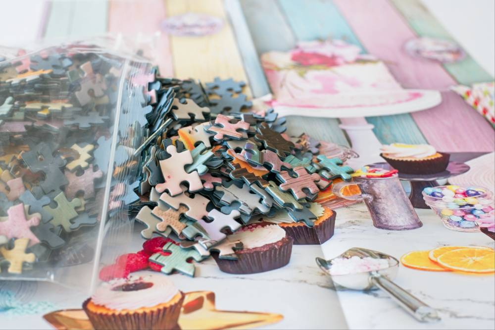 A pile of small jigsaw puzzle pieces spilled from a clear plastic bag onto the poster, which shows cupcakes, desserts, and colorful pastel background panels.