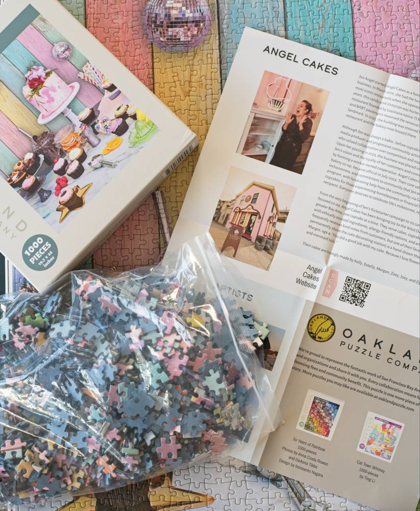 A 1000-piece jigsaw puzzle set from Oakland Puzzle Company, including the illustrated puzzle box, a clear resealable bag of loose puzzle pieces, and an open poster titled 'Angel Cakes' on the completed puzzle.