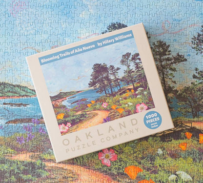 A boxed jigsaw puzzle from Oakland Puzzle Company titled 'Blooming Trails of Año Nuevo' by Hilary Williams, placed on top of a completed puzzle. The box shows a colorful landscape illustration with trees, water, flowers, and a path, plus a blue badge that says '1000 PIECES'.