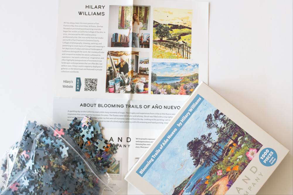 A jigsaw puzzle box titled 'Blooming Trails of Año Nuevo' by Hilary Williams, shown with loose puzzle pieces and a poster with 'About' details on a white background.