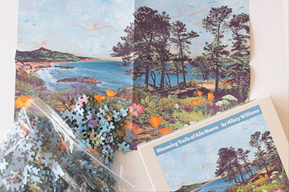 A jigsaw puzzle set for 'Blooming Trails of Año Nuevo' by Hilary Williams, showing the printed cover, a clear bag of loose puzzle pieces, and a partially assembled portion of the coastal landscape puzzle on a white background.