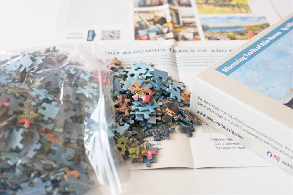 Open jigsaw puzzle box and loose puzzle pieces spread on the poster. The box shows the Oakland Puzzle Company brand.