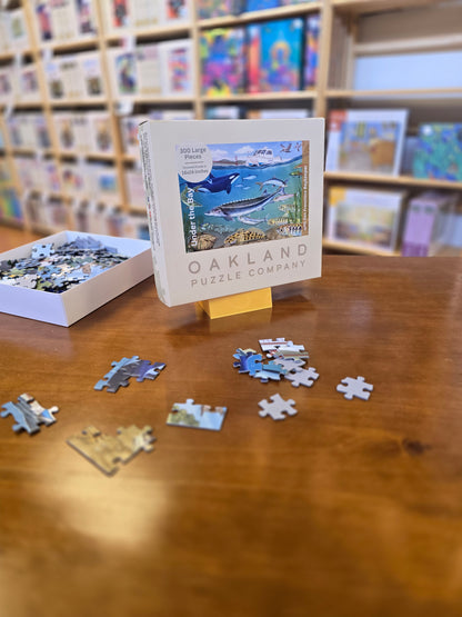 Oakland Puzzle Company puzzle box propped in a yellow box stand with scattered pieces on a wooden surface