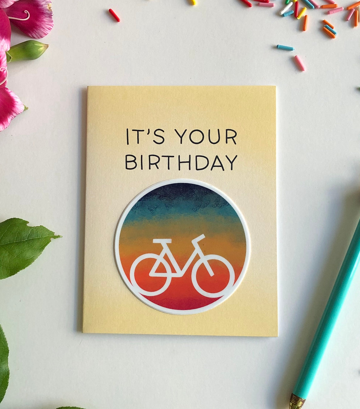 Photo of a greeting card that says "It's Your Birthday with a colorful gradient sticker with the white silhouette of a bicycle.