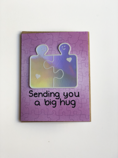 A greeting card with a jigsaw puzzle pattern and the text Sending you a big hug with a sticker of two interlocked puzzle pieces with smiley faces and hearts